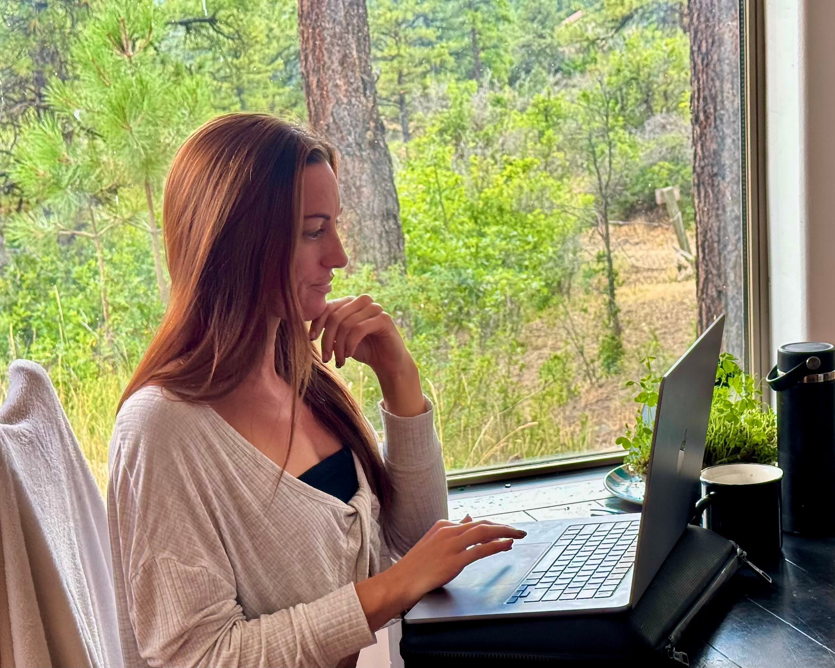 Alyssa Arroyo in profile at a laptop, chin resting on her hand, seated beside a tall window looking out on a sunlit pine forest.