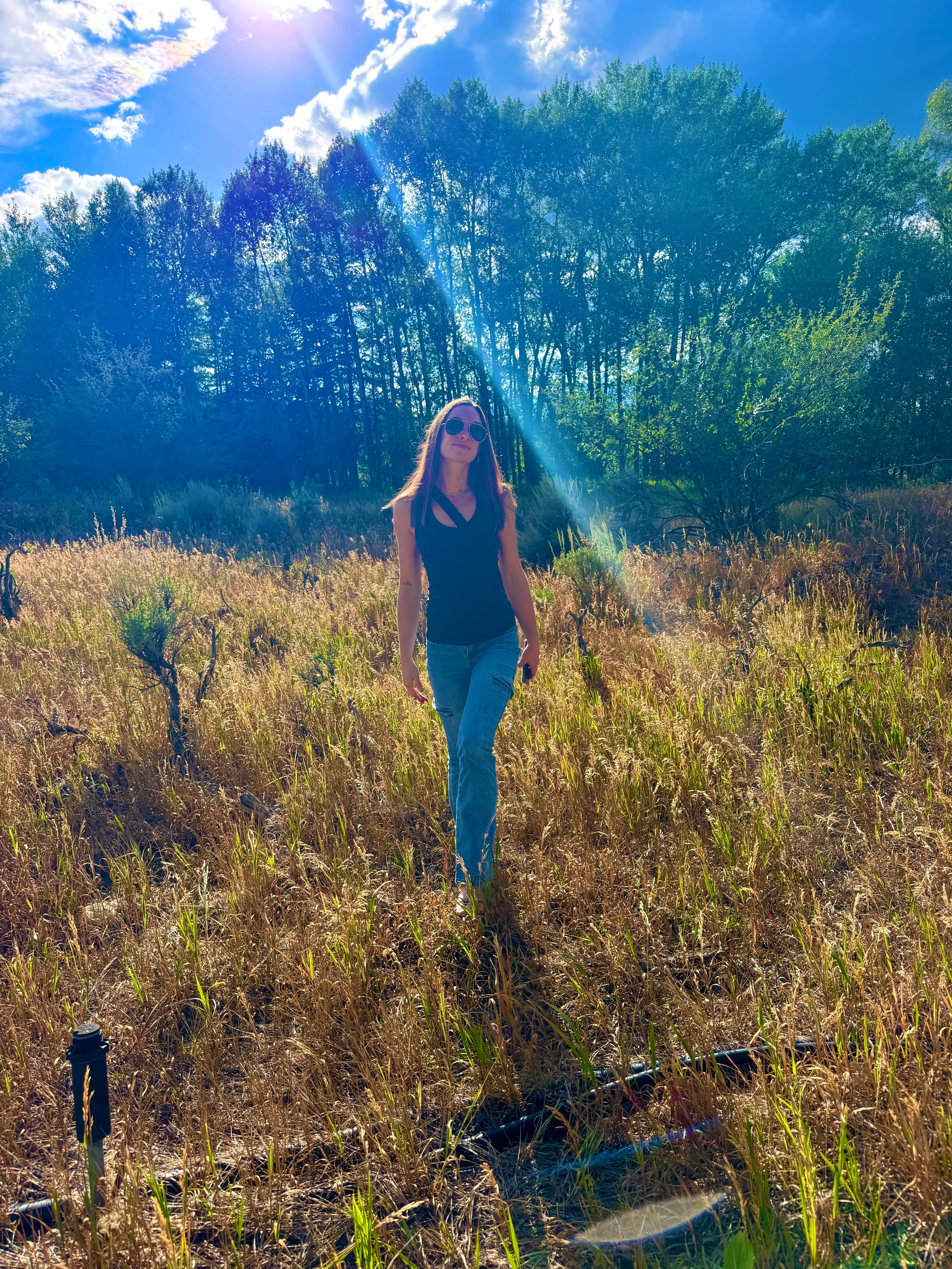 Alyssa Arroyo walking through a sunlit meadow of tall golden grass, wearing sunglasses and a black top, with a stand of aspens behind her under a partly cloudy blue sky.