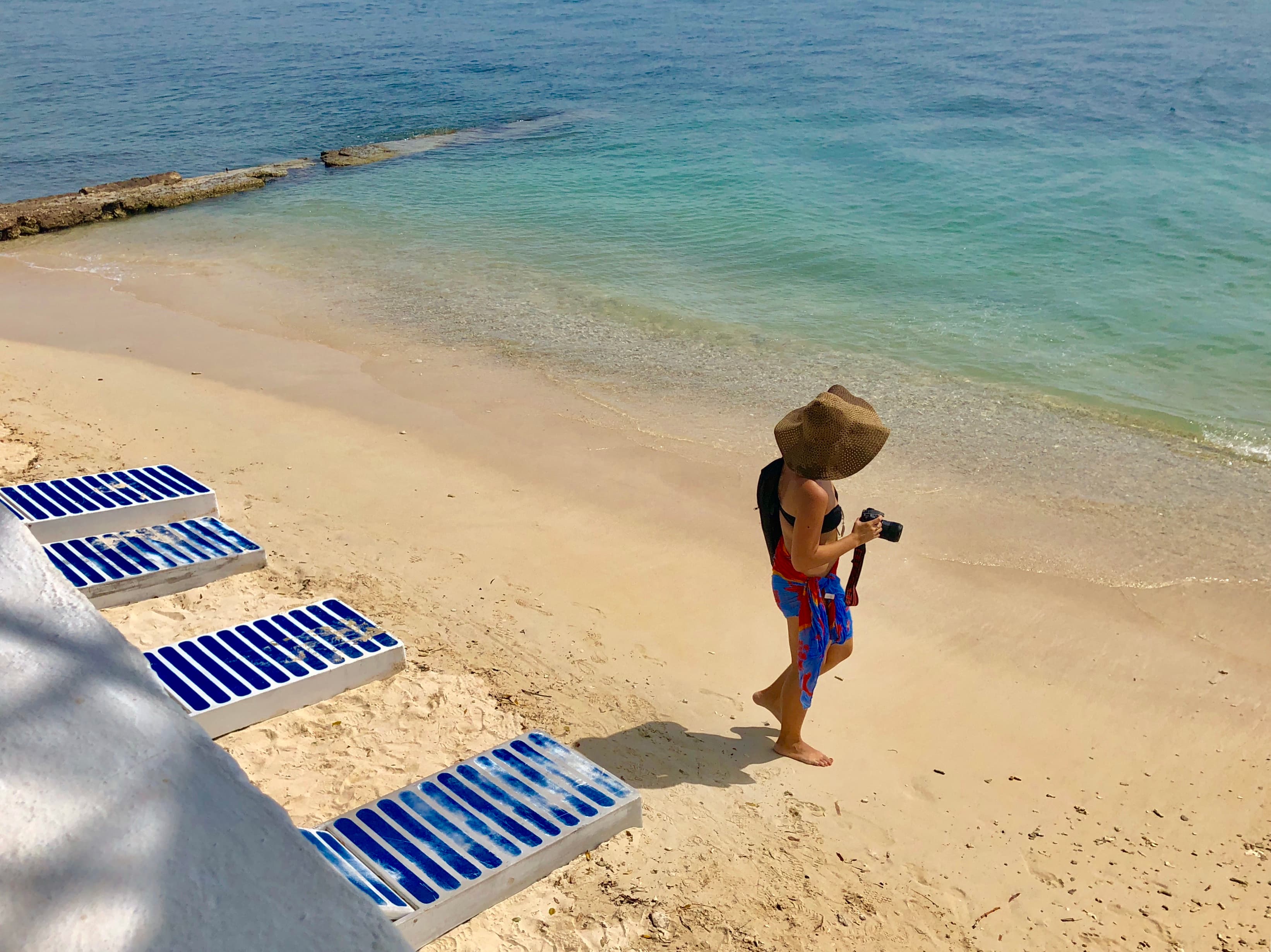 A woman in a sun hat walking a beach at Cocoliso, camera in hand, with turquoise water and a stone breakwater behind her.