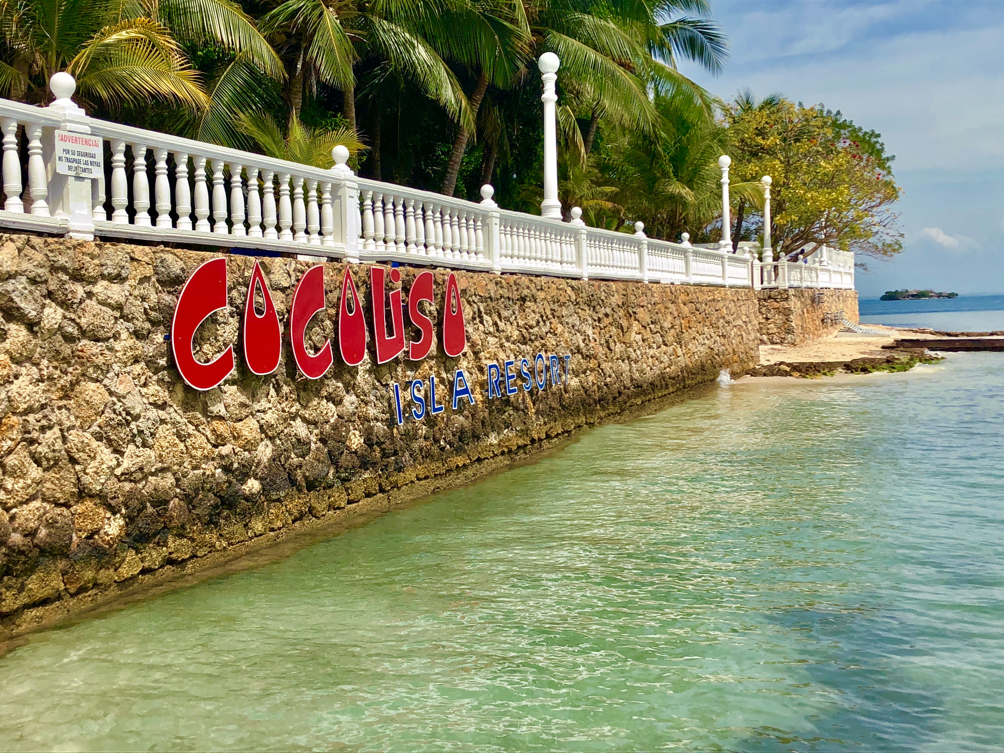 The Cocoliso Isla Resort sign set into a stone seawall lined with a white balustrade, palm trees and clear shallow water along the shore.