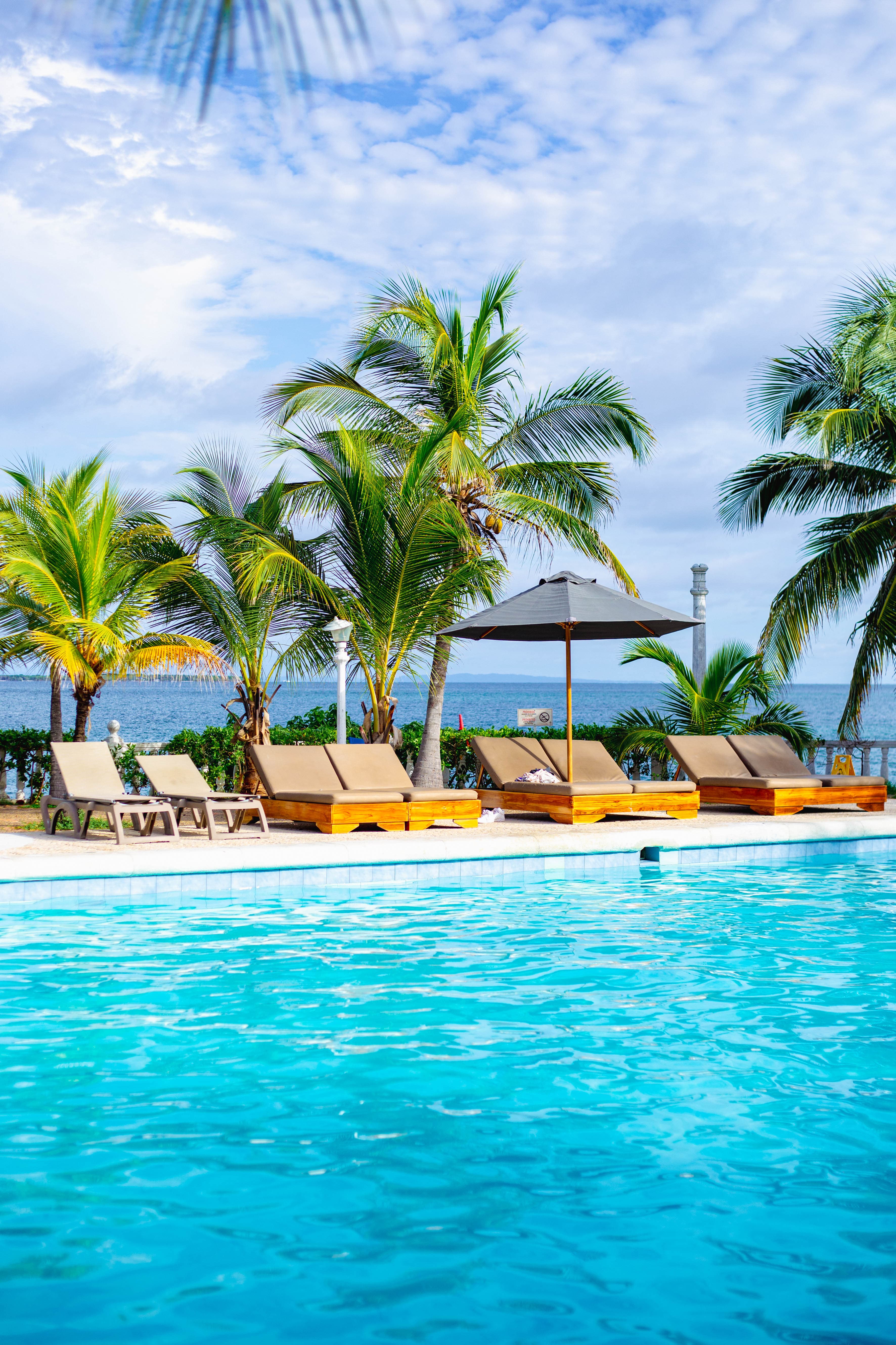 Wooden lounge chairs and a shade umbrella along the edge of a turquoise pool at Cocoliso, palm trees and the open sea behind them.