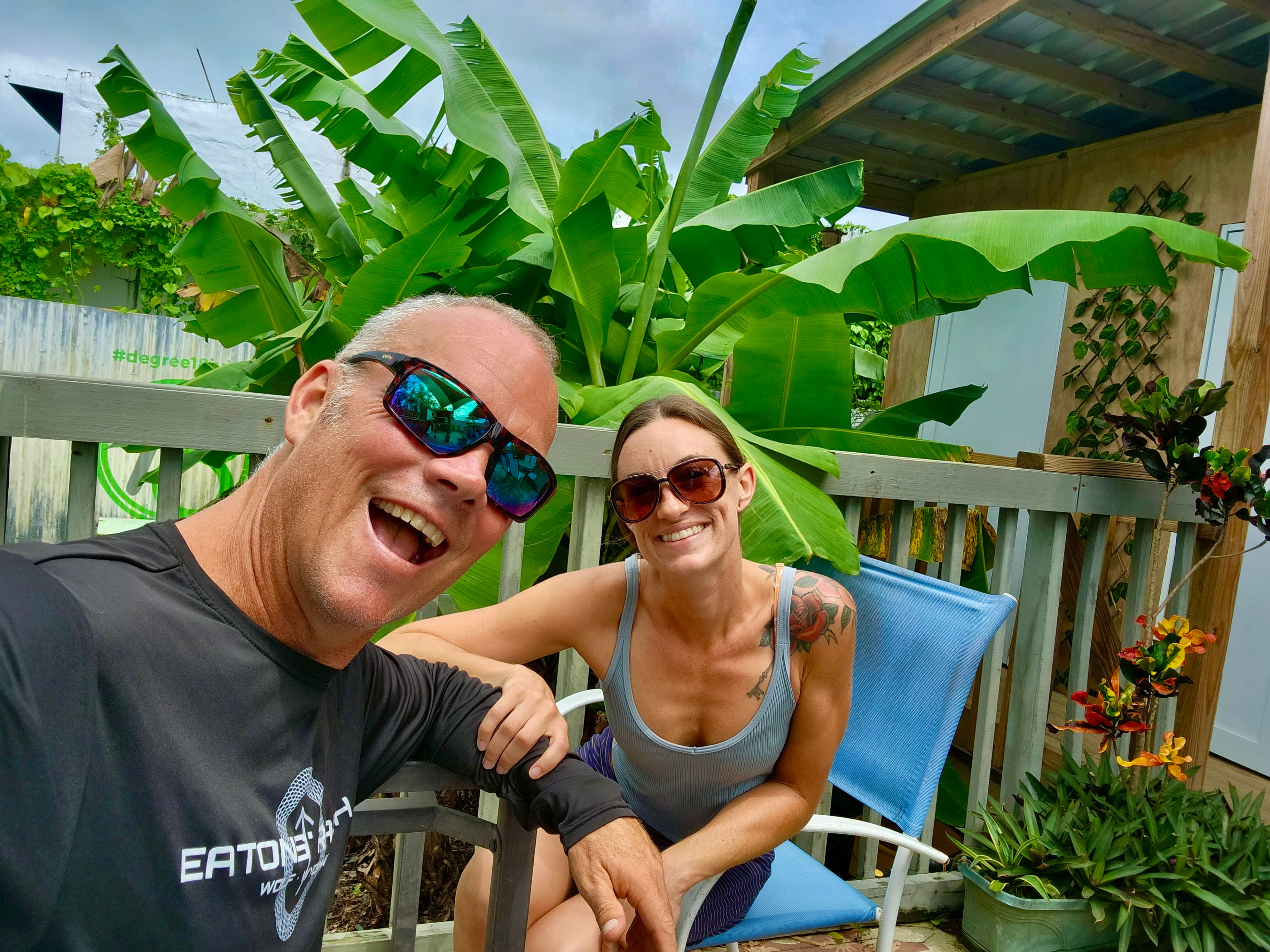 Holland Duell and Alyssa Arroyo laughing together on a shaded deck, both in sunglasses, with banana palms and a timber pavilion behind them.