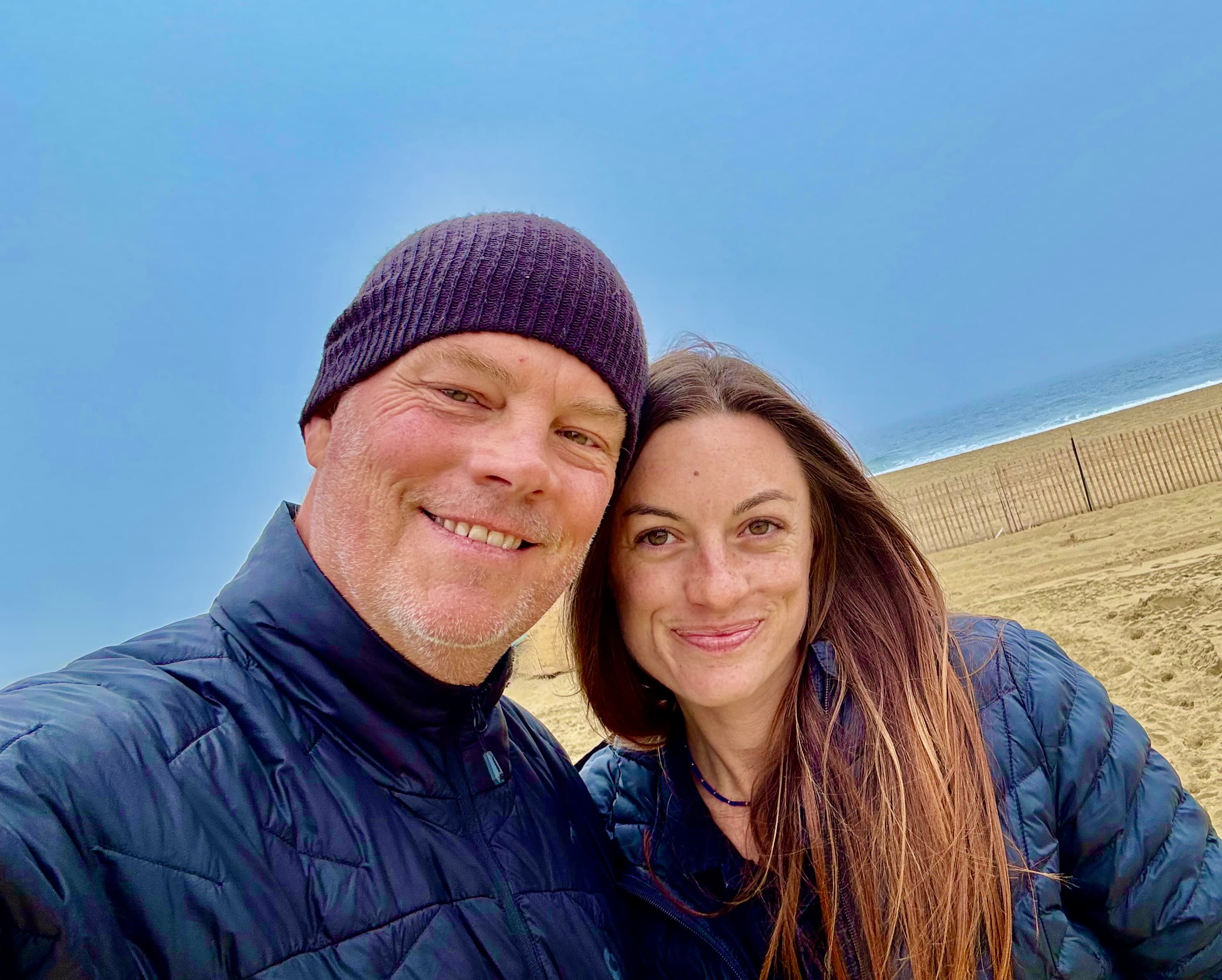 Holland Duell and Alyssa Arroyo cheek to cheek and smiling toward the camera on a beach under a clear blue sky, both in navy jackets and Holland in a knit beanie, with a dune fence and the surf behind them.