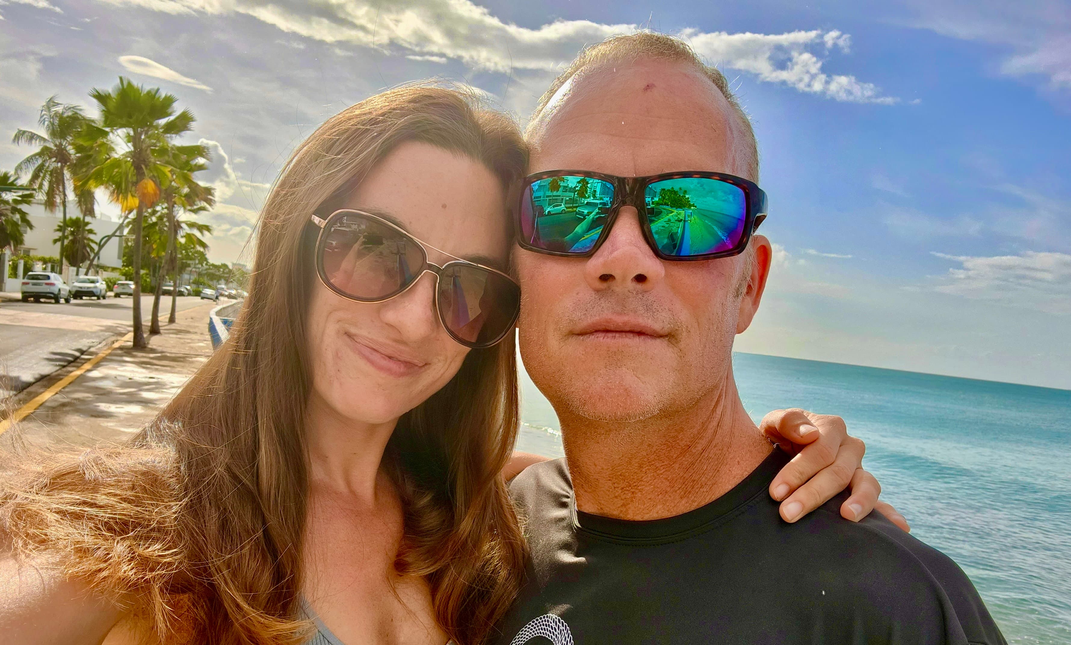 Holland Duell and Alyssa Arroyo side by side on a seafront promenade, both wearing sunglasses, with palm trees and the ocean behind them.