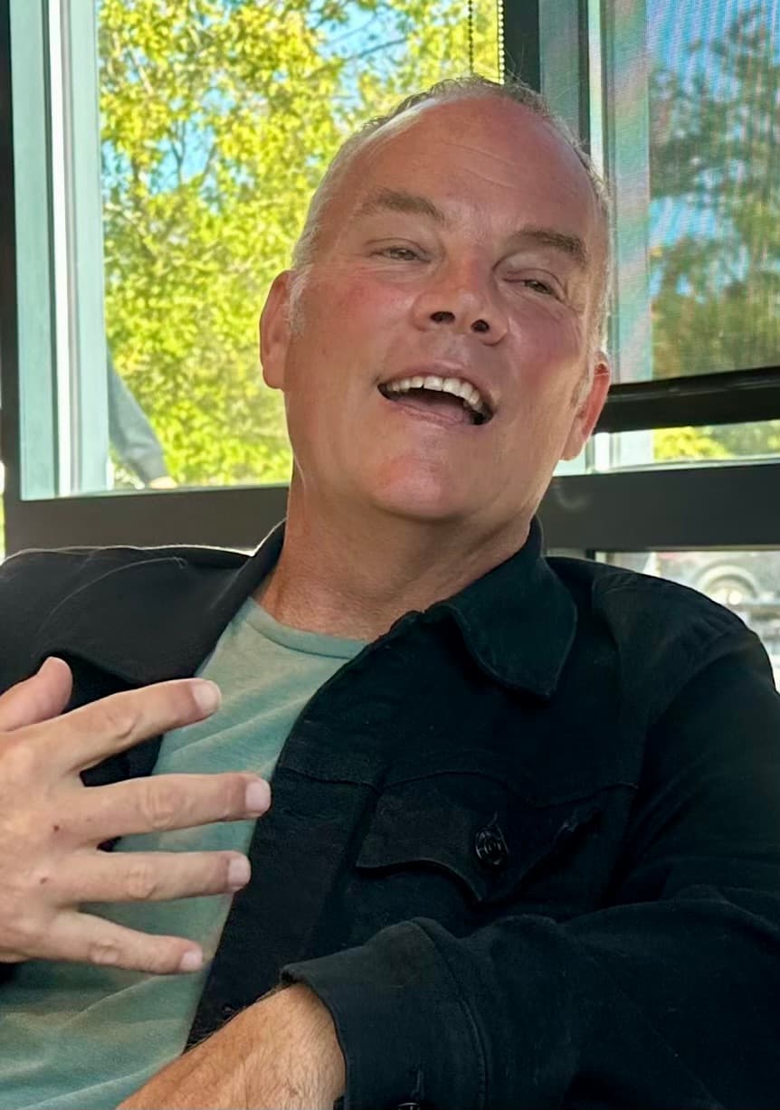 Holland Duell laughing mid-conversation, seated beside a sunlit window in a dark jacket over a grey T-shirt.