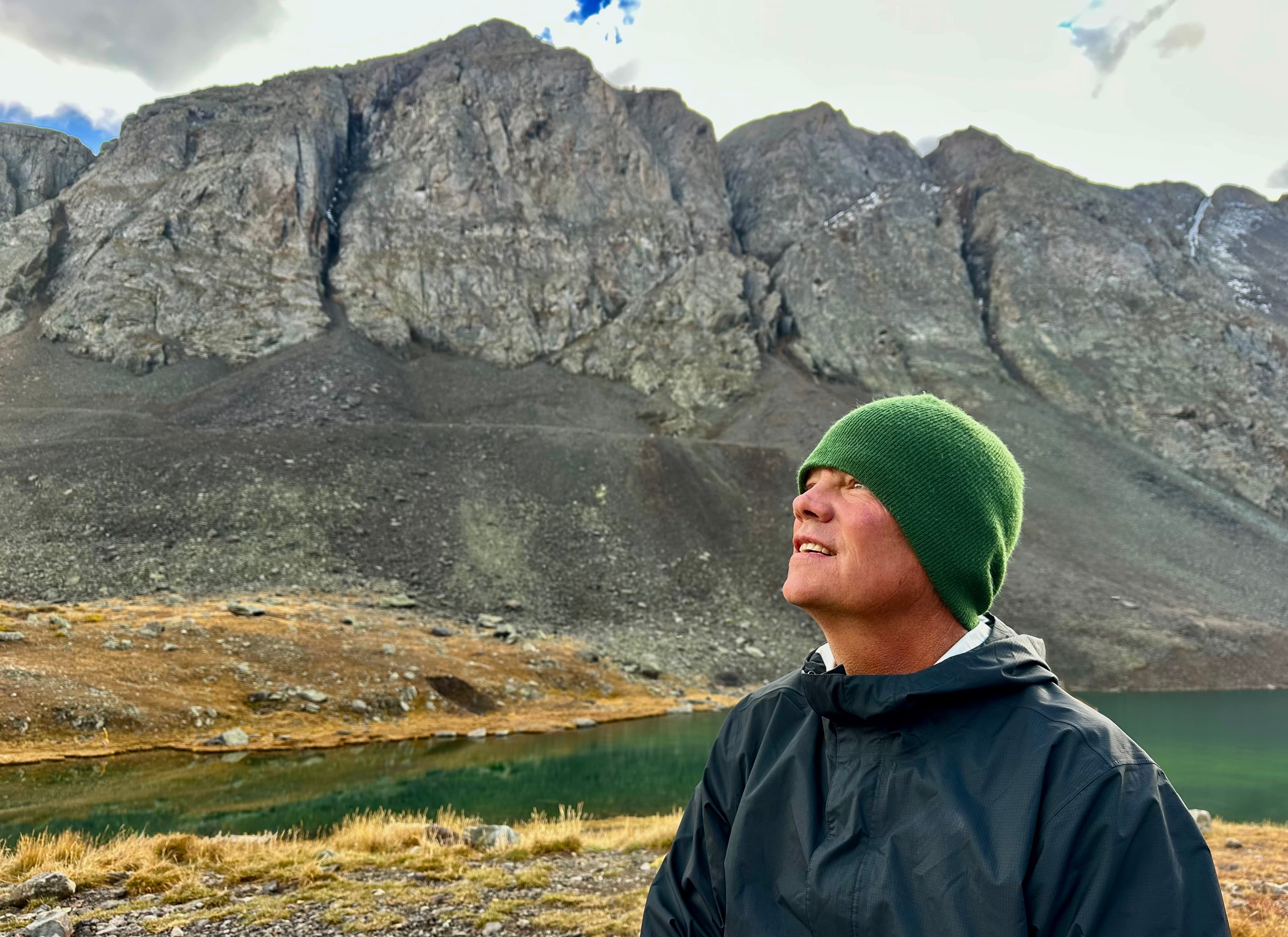 Holland Duell, in a green knit beanie and dark rain jacket, standing beside an alpine lake and looking up toward the towering rock face behind him.