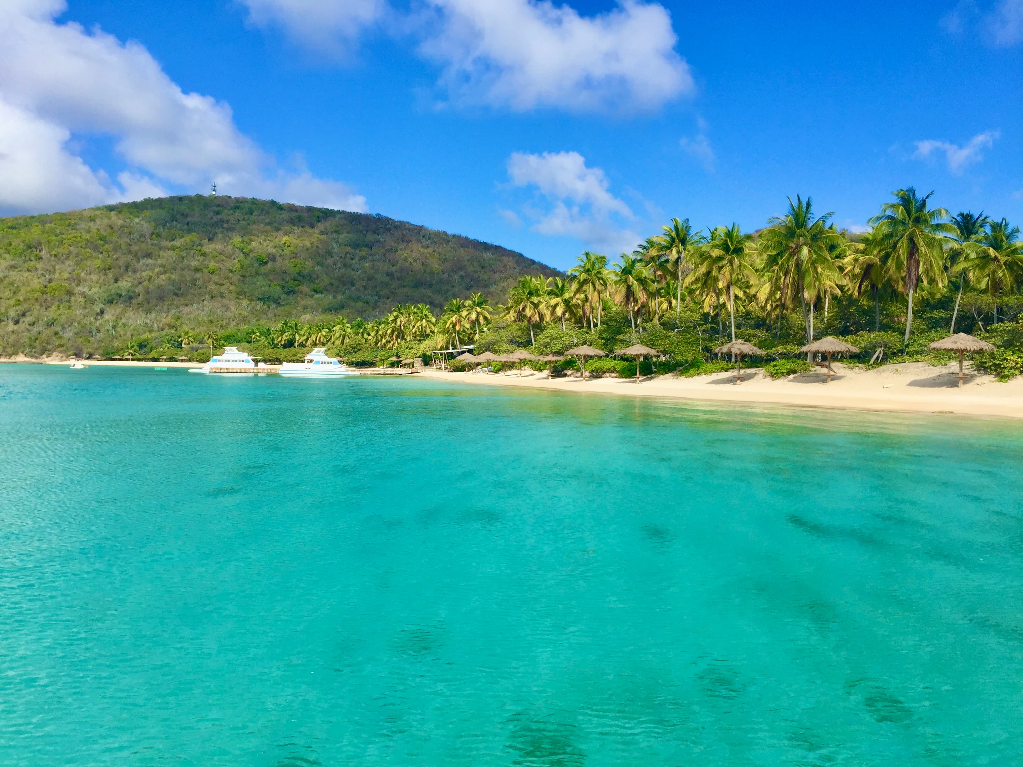 A sheltered turquoise bay, with thatched sun shades and palms along a pale sand beach and a forested hill rising behind.