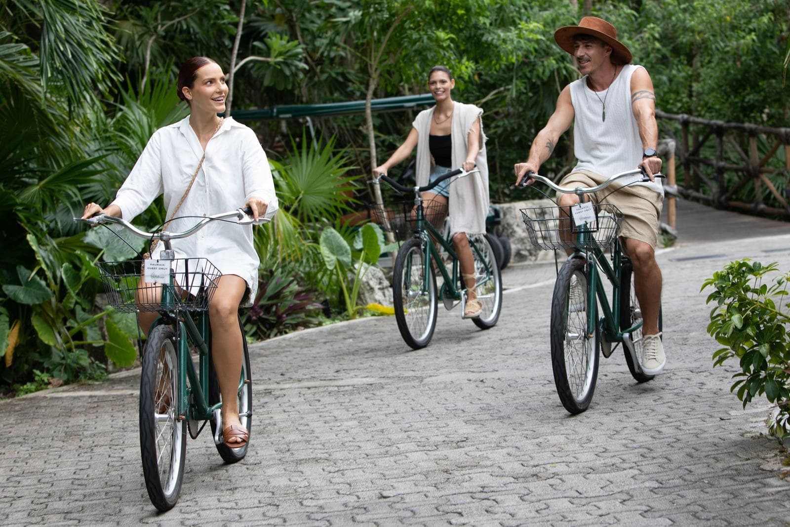 Two women and a man riding green bicycles along a paved path through the jungle, the woman in front smiling back over her shoulder