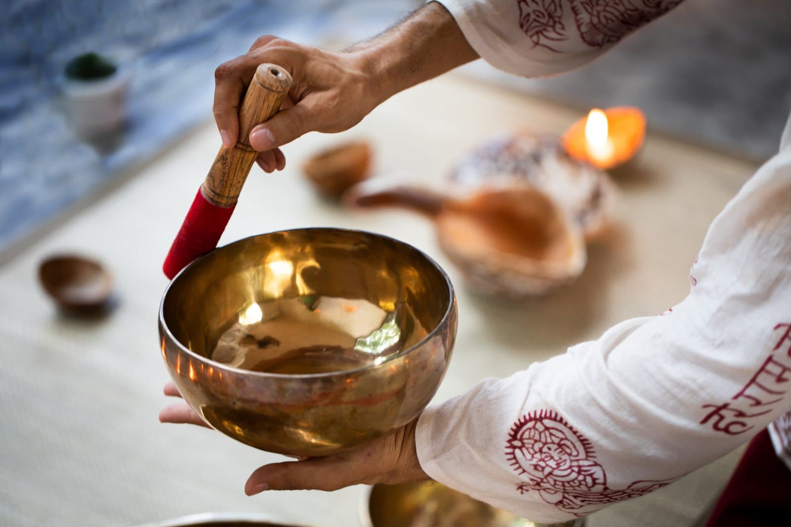 Hands holding a brass singing bowl and a wooden mallet, with a lit candle and shells softly out of focus behind