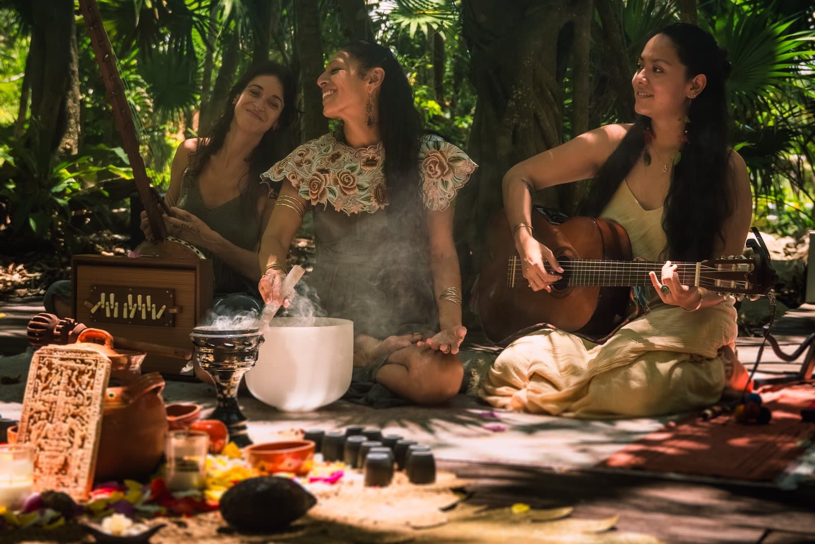 Three women making music on a jungle deck, one strumming a guitar, one with a crystal singing bowl and rising incense smoke, one with a stringed instrument, around an offering of petals, candles and a cacao pod