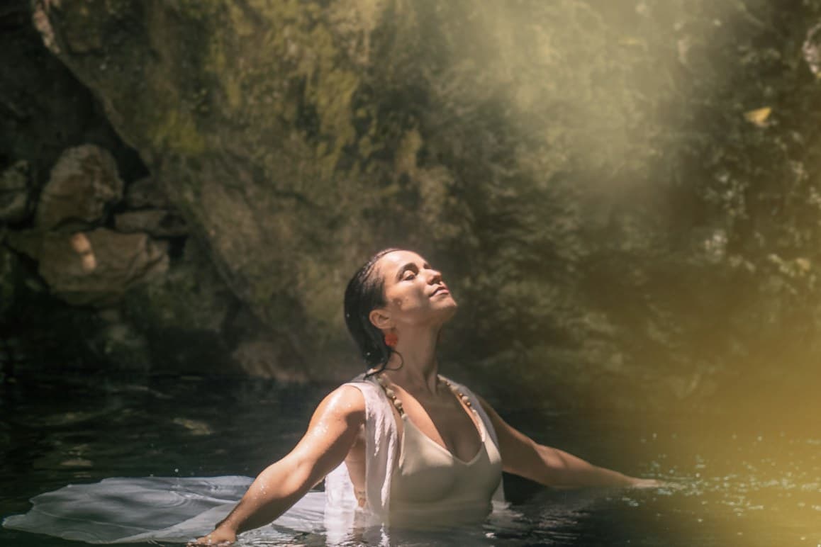 A woman standing in the clear water of a cenote with her head tipped back and eyes closed in a beam of sunlight, mossy rock rising behind her