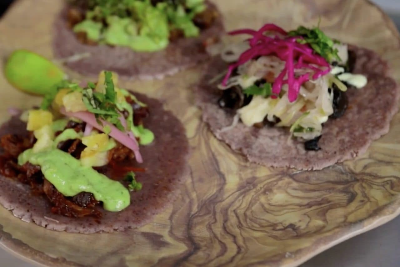 Three tacos on blue corn tortillas topped with pineapple, pickled red cabbage and green sauce, served on a wooden plate