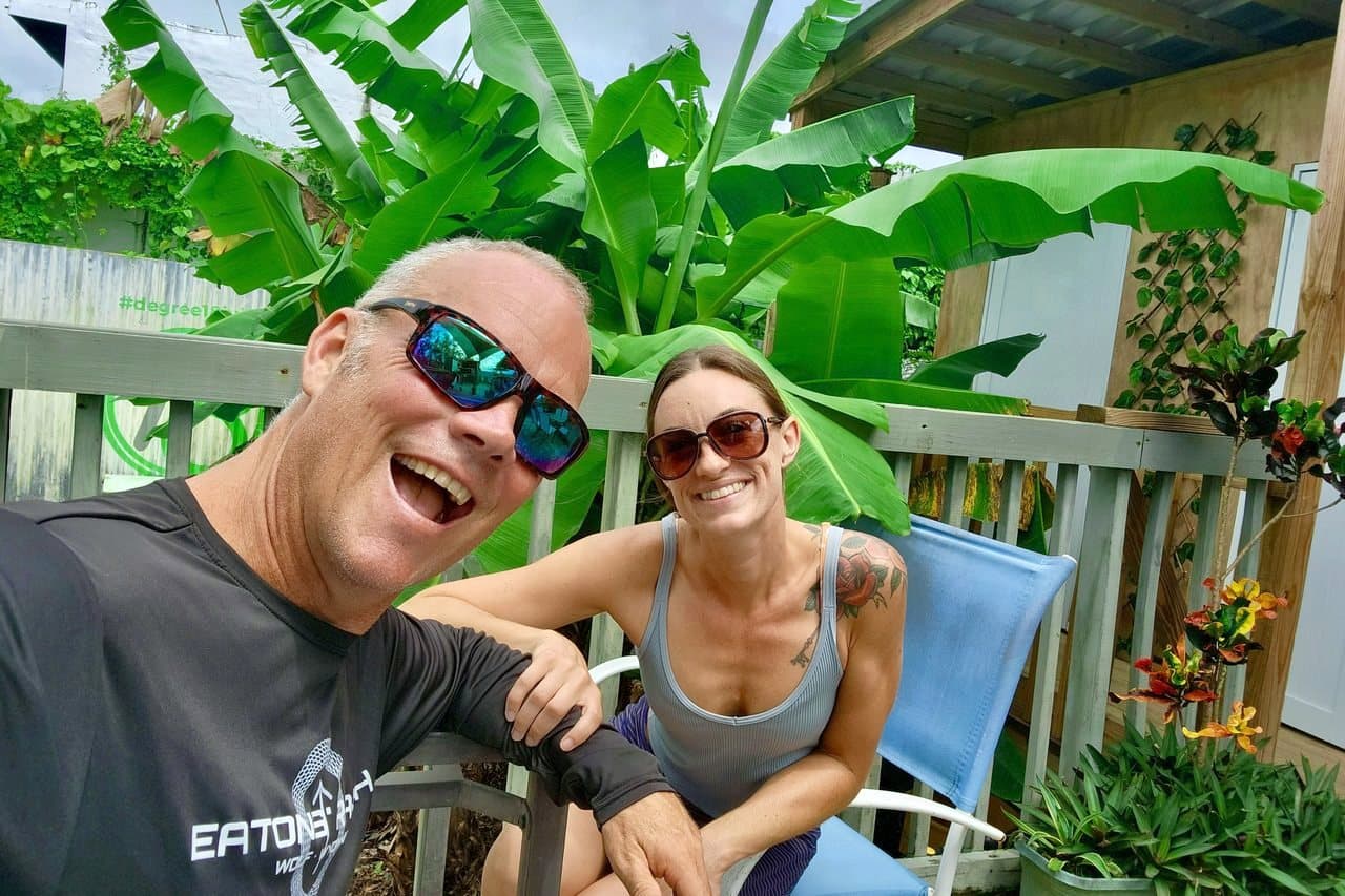 Holland Duell and Alyssa Arroyo smiling on a wooden deck in front of banana plants, Holland in sunglasses leaning toward the camera