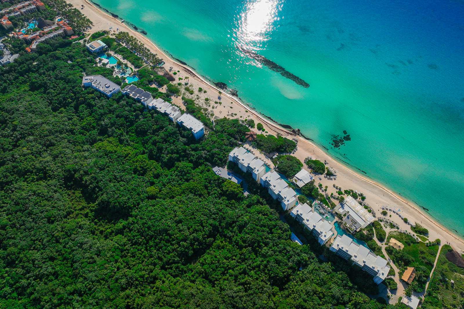 An aerial view looking straight down on Palmaïa's white buildings stepping along a white-sand beach, with turquoise pools between them, dense jungle behind and the turquoise Caribbean beside