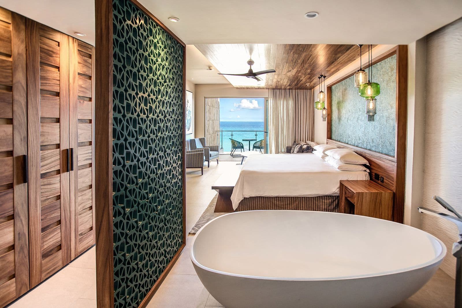 King Suite with Bathtub: a freestanding white bathtub in the foreground and a king bed under a wood ceiling, with the sea visible through the balcony doors