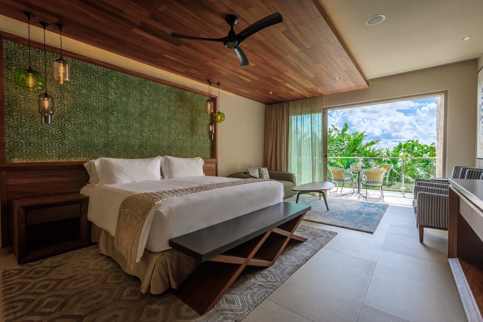King Suite: a king bed against a green patterned wall under a wood ceiling, with a sofa and balcony doors opening onto palms and jungle