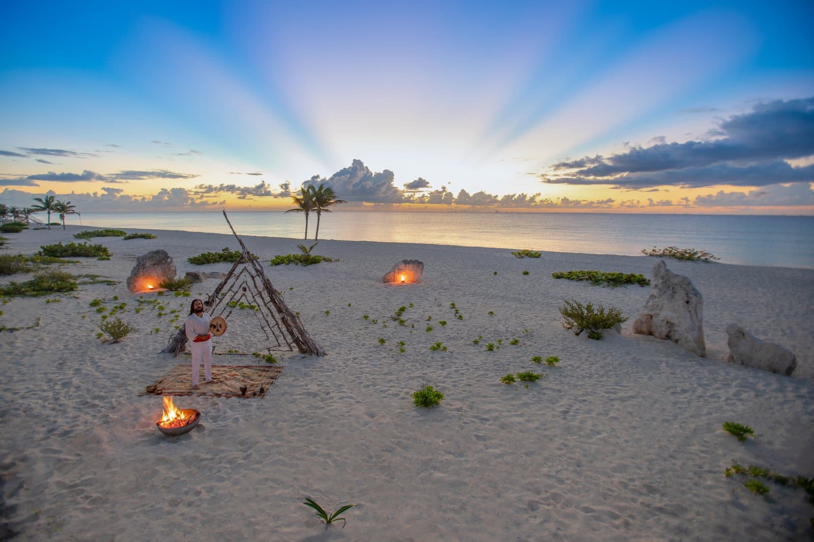 A man in white holding a round drum beside a driftwood teepee and a bowl of fire on a wide, empty white-sand beach at dawn, with rays of light fanning across the sky above the sea