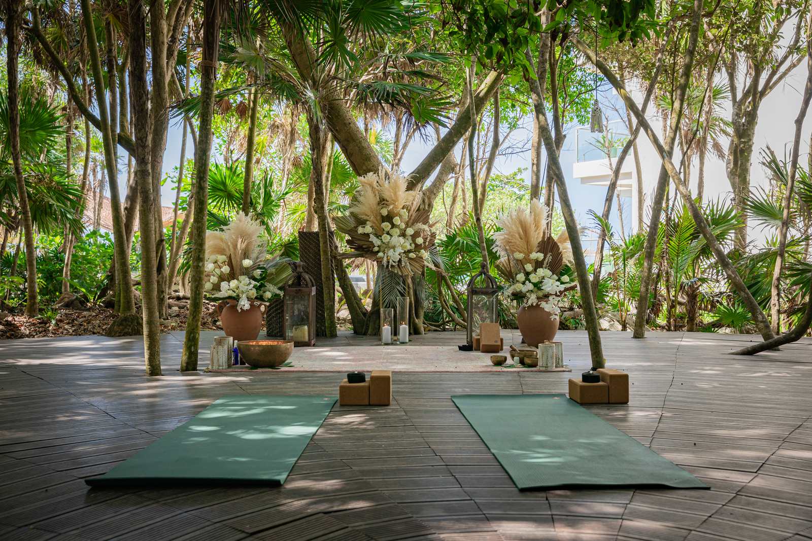 A jungle clearing of tall, slender trees with a wooden deck set for practice: two green yoga mats, cork blocks, a singing bowl and large arrangements of white flowers and pampas grass