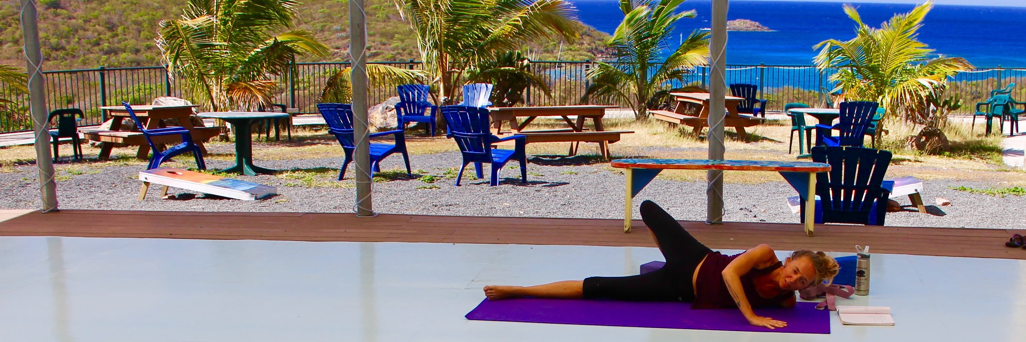 A woman stretching on a purple yoga mat on an open-air deck, with Adirondack chairs, picnic tables, palms and the sea beyond.