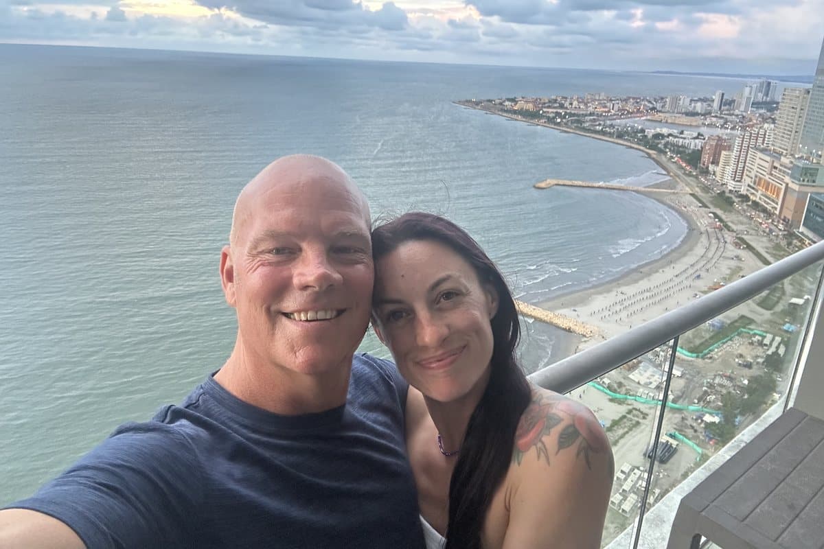Holland Duell and Alyssa Arroyo smiling cheek to cheek in a selfie on a high balcony, with a curving coastline, a sandy beach and a city on the shore behind them under a cloudy dusk sky.